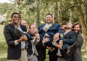 A group of groomsmen laugh together while posing for casual photos outdoors in a wooded area.