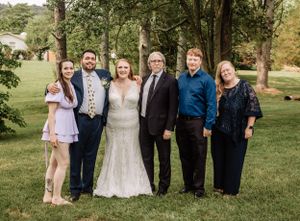 Large wedding party assembles for group photos on a green lawn with mature trees.