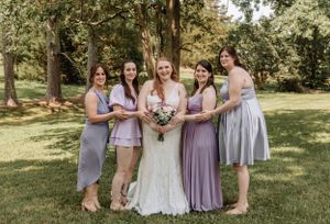 Wedding party in purple and gray dresses poses joyfully outdoors in a park setting.