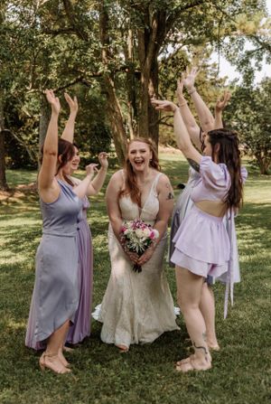 Bridesmaids in lavender dresses celebrate with energetic poses in park setting.