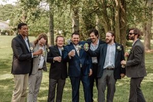 Groomsmen in mixed formal attire pose playfully for candid shots in a wooded setting.