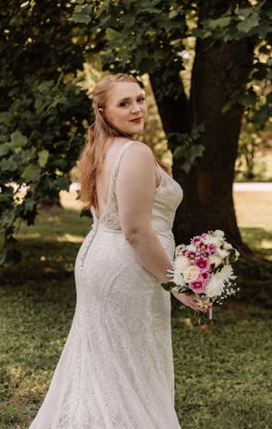 Plus size bride wearing a detailed lace wedding gown poses with flowers in a park setting.