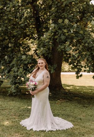 A bride in a lace wedding dress poses with a pink bouquet under a shady tree.