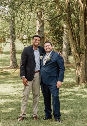 Two wedding guests smile together while standing on grass in a wooded outdoor setting.
