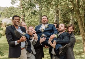 Groomsmen pose playfully for candid photos during an outdoor celebration among trees.