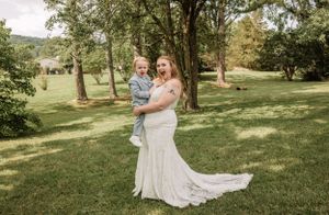 A bride in white lace gown holds a small child in a sunny outdoor setting.