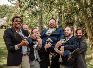 Wedding party members share candid laughs while standing together under trees at an outdoor venue.