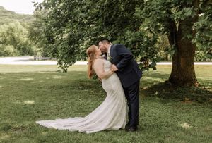 A couple in wedding attire shares a romantic moment under a shady tree on a sunny day.