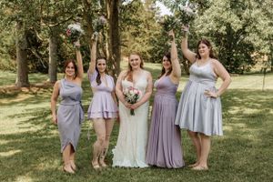 Wedding party wearing coordinated purple and gray attire stands in formation on sunny lawn.