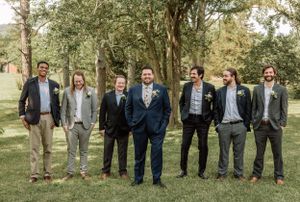 Groomsmen in varied suits pose together on grassy lawn with trees in background.