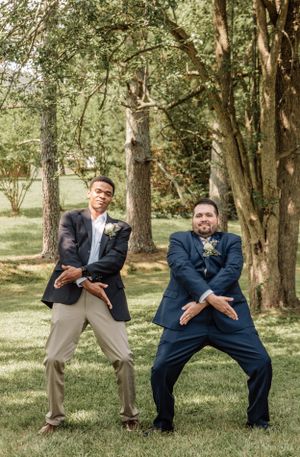 Two wedding guests strike playful poses while standing on grass in a wooded outdoor setting.