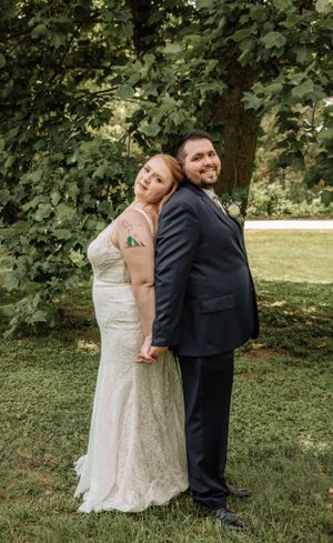 Playful wedding couple poses back-to-back in outdoor garden setting wearing formal attire.