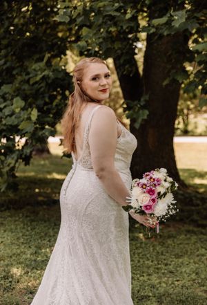 Bride in a sleeveless lace wedding dress holds a pink and white bouquet in an outdoor garden setting.