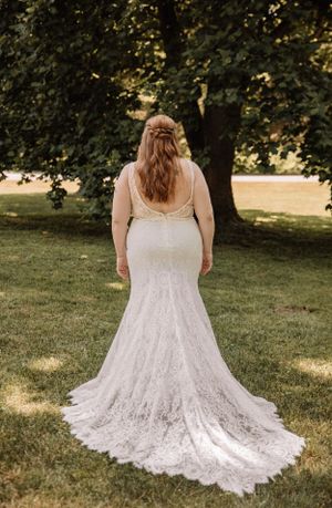 A plus-size wedding dress with intricate lace detailing and straps photographed from behind in a park setting.