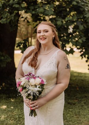 A person in a white wedding dress smiles while holding a pink and white bouquet against green foliage.