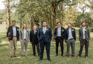 Wedding party members stand in casual formation on grass with trees behind them.