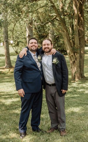 Three wedding party members stand together smiling in a sunlit wooded area.