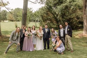 Playful wedding party poses informally on grass with trees in background.