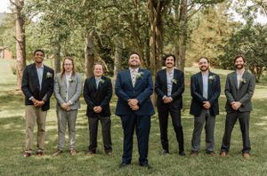 Wedding party members in navy and gray suits pose for formal group photo outdoors.
