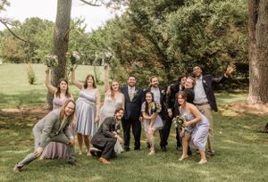 Wedding party members strike fun poses on grass beneath towering trees.