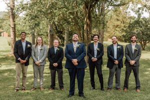 Wedding party members in formal attire pose in line formation under trees.