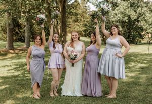 Bridal party members in lavender and gray dresses gather for outdoor photos in a park setting.