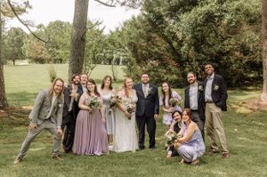Wedding party members strike relaxed poses on lawn beneath towering pine trees.