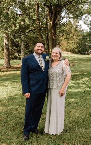 Couple in formal attire poses together on grass with trees in background.