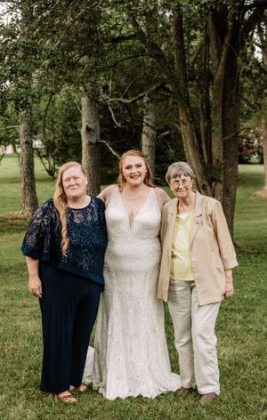 Wedding party poses outdoors with bride in white lace dress and guests in navy and neutral tones under trees.