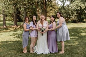 Bridal party wearing lavender and gray dresses stands together on grassy lawn.