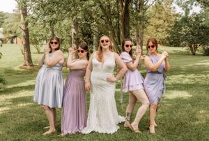Wedding party members in soft pastel dresses gather for photos on a grassy hillside.