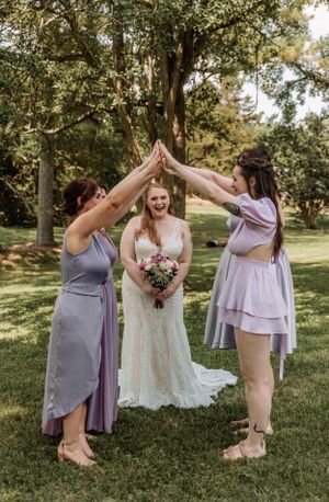 Wedding party members in purple tones create playful high-five poses on sunny lawn.