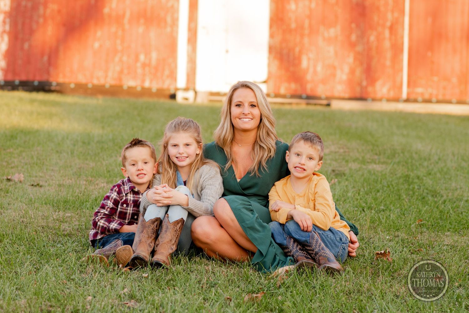 Family seated together on grass with red barn in background during golden hour photography session.