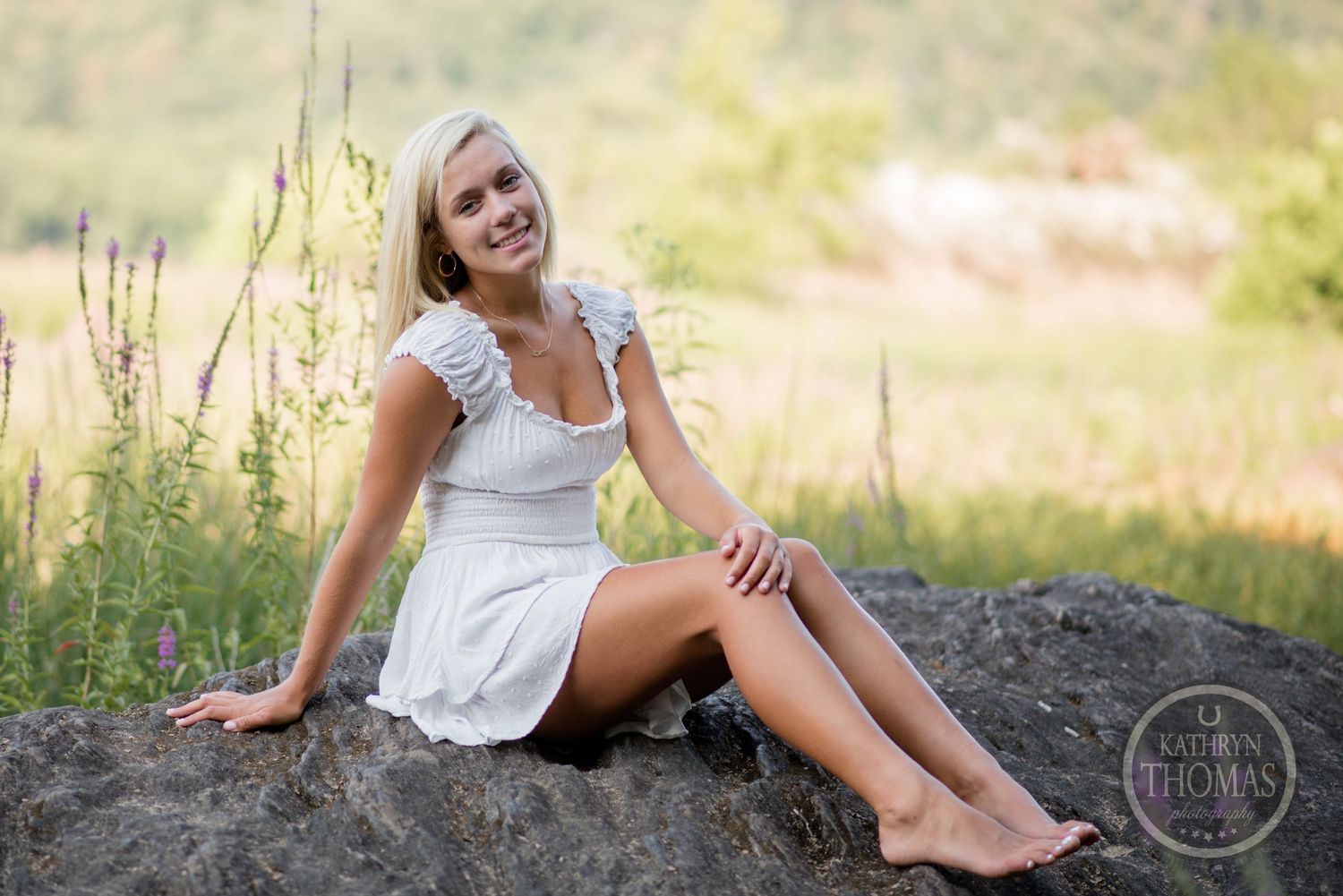 A summer portrait taken outdoors shows someone in a white sundress sitting on a rock in a natural setting.
