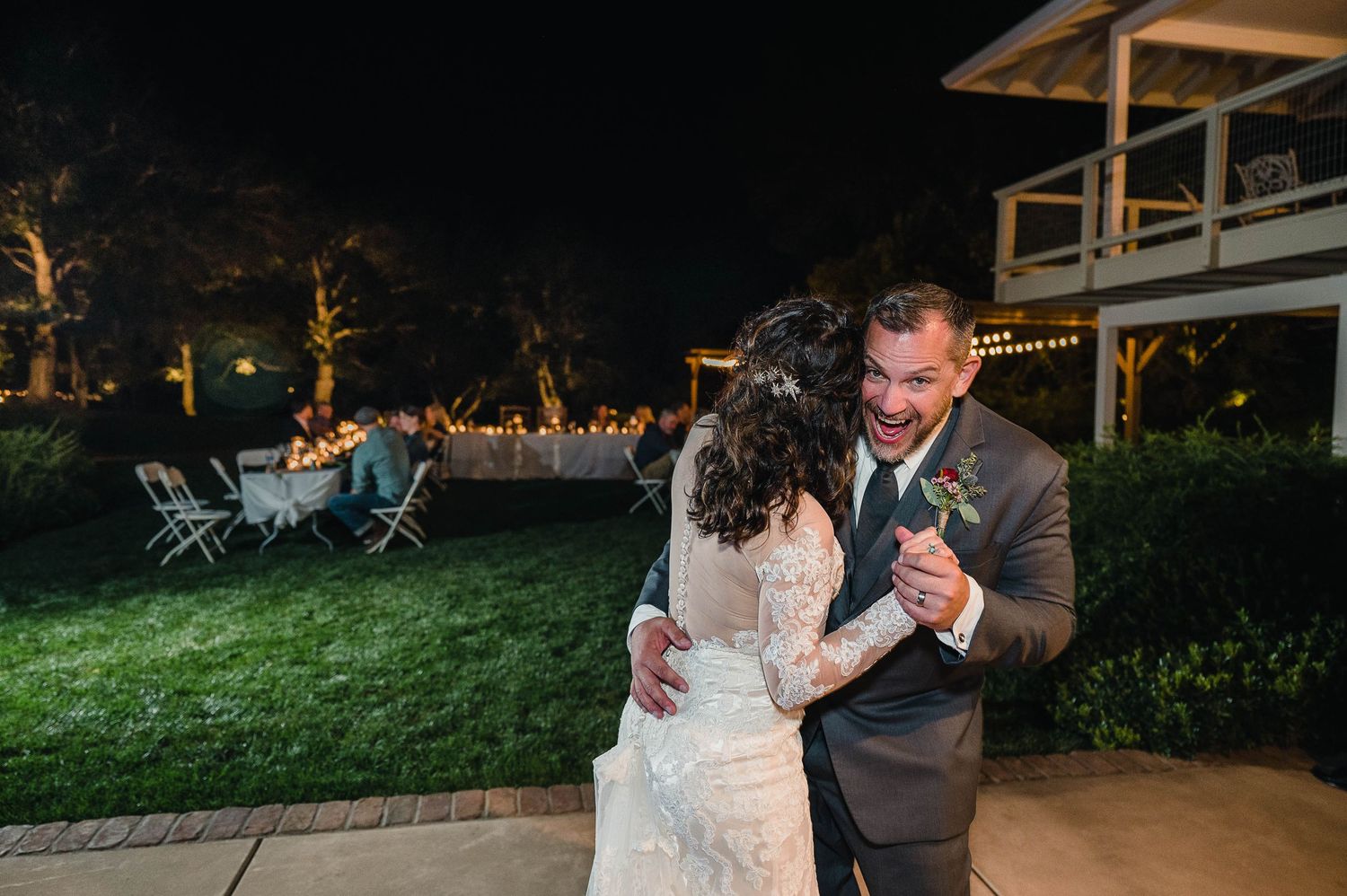 Lace-dressed couple dancing together on lit garden lawn during evening wedding celebration.