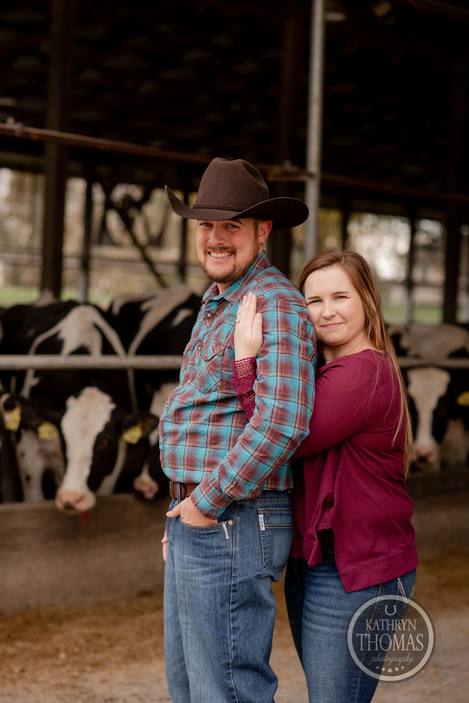 Happy couple poses together in front of dairy cow stalls wearing casual western attire.
