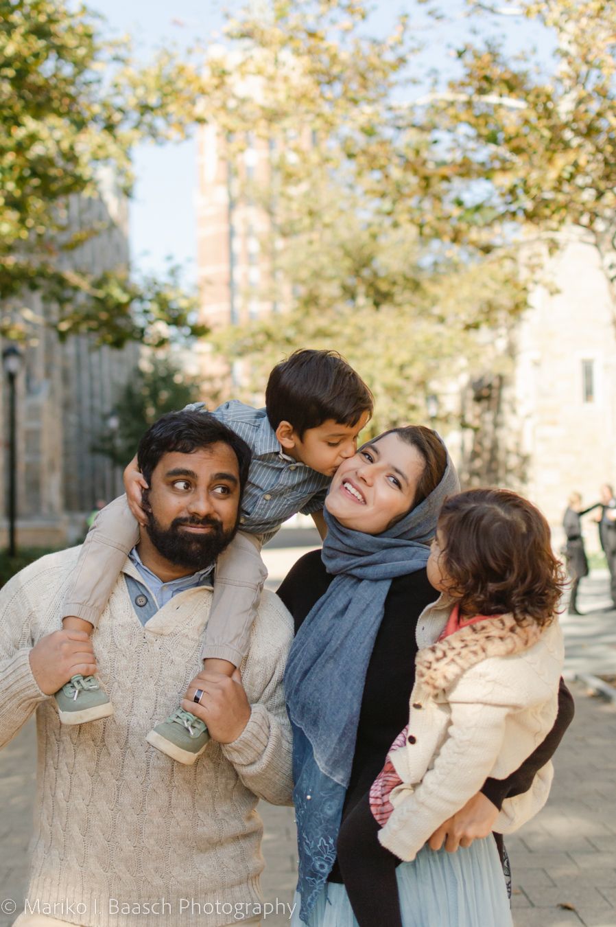 Happy family of four with two young children outdoors in a sunny urban park setting.