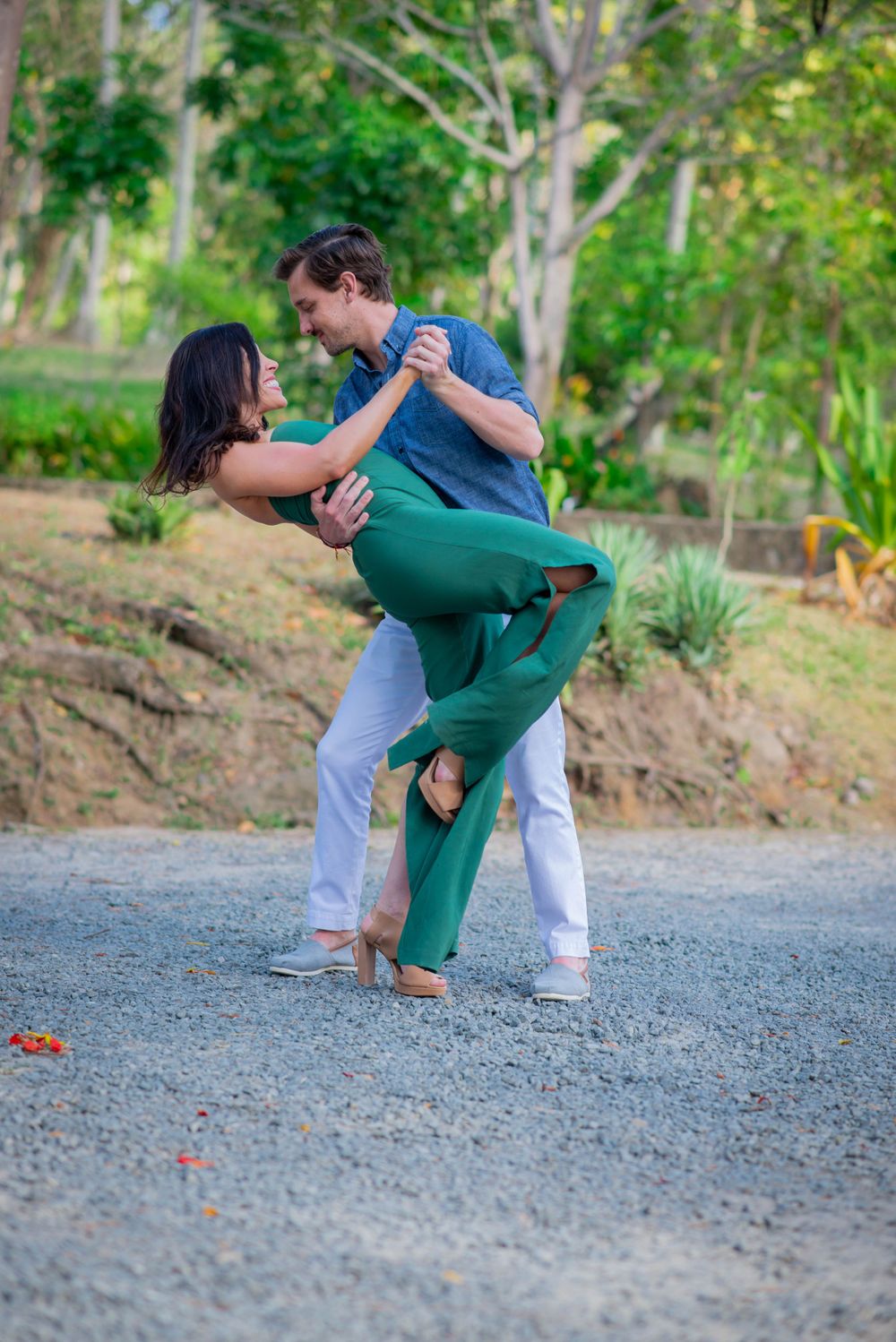 Couple in casual and formal wear dancing playfully on gravel path surrounded by greenery.
