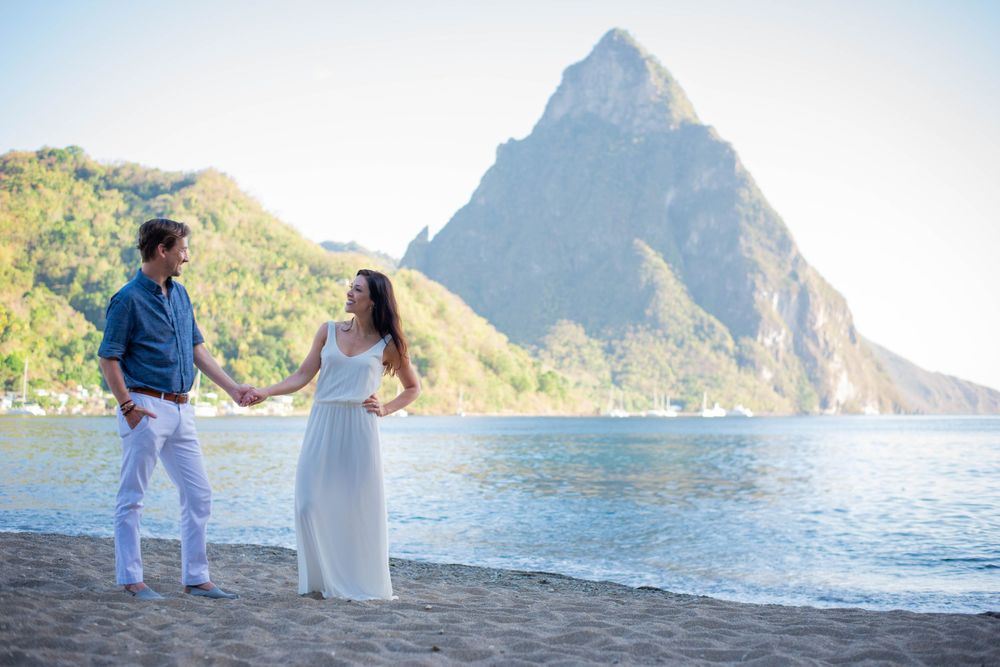 Couple holding hands on sandy beach with Saint Lucia's volcanic peaks in distance.
