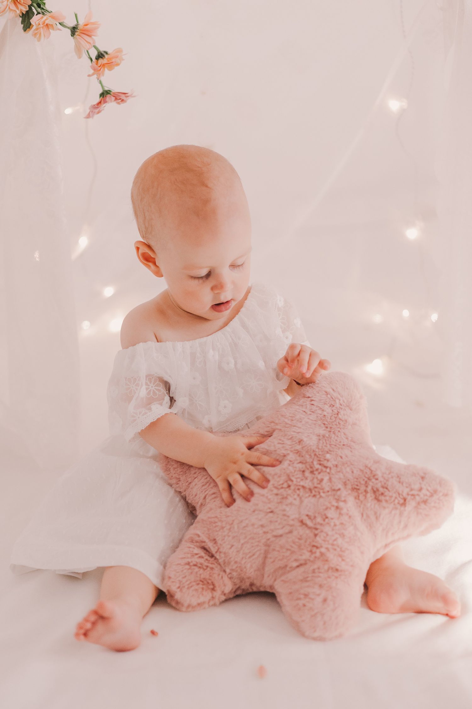 Baby in white dress playing with pink plush star toy on light background.
