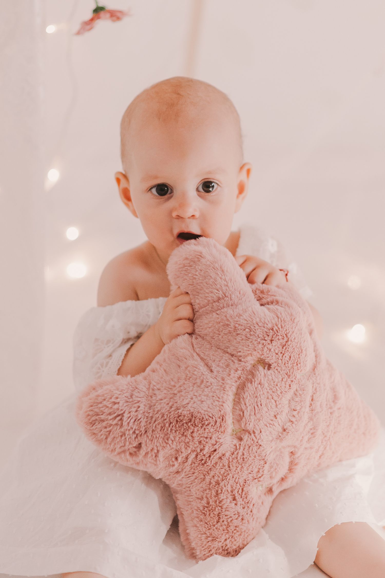 Baby in white dress holding pink plush toy against dreamy light background.