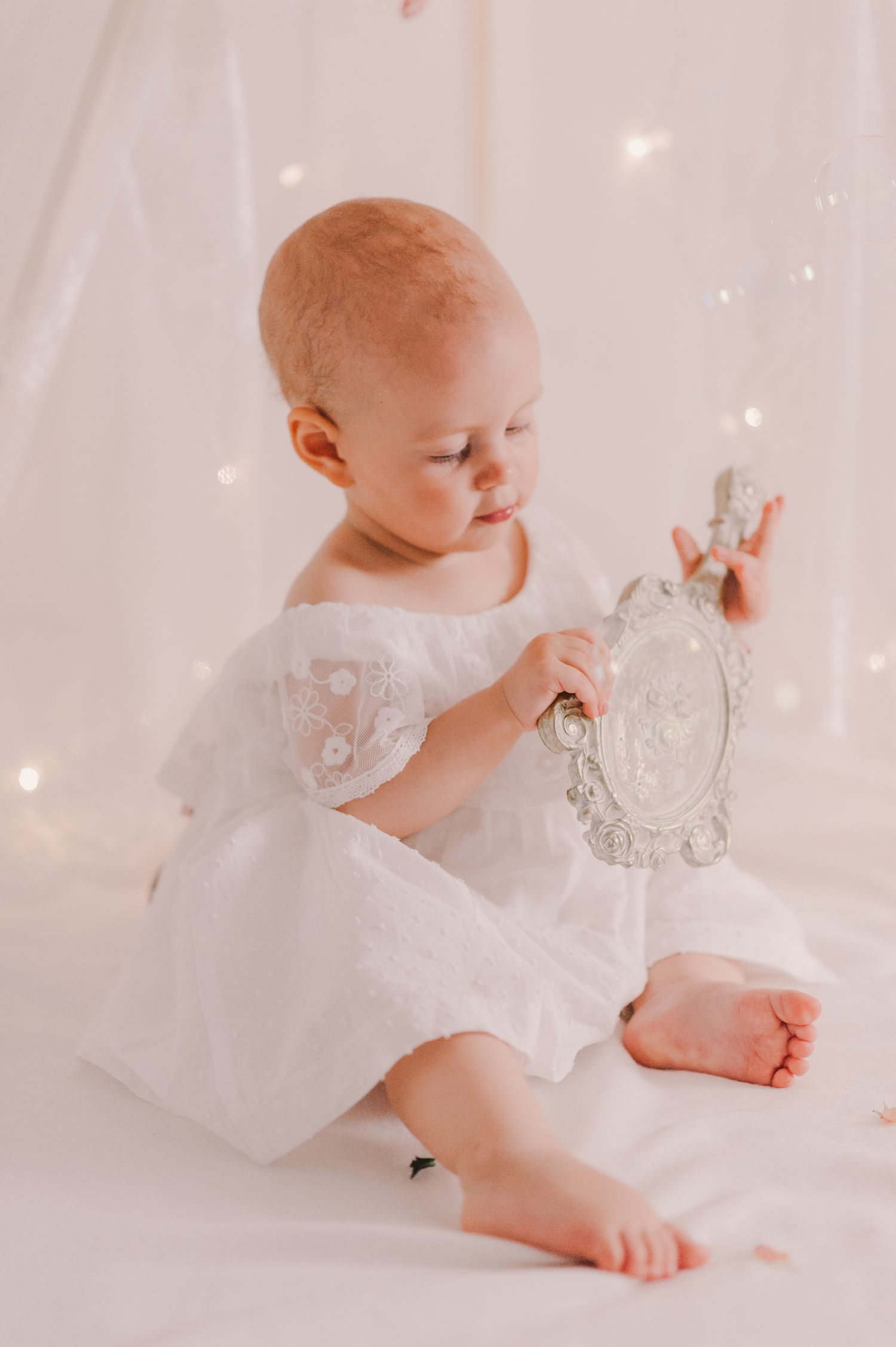 Baby in white dress holding ornate vintage mirror while sitting on light background.
