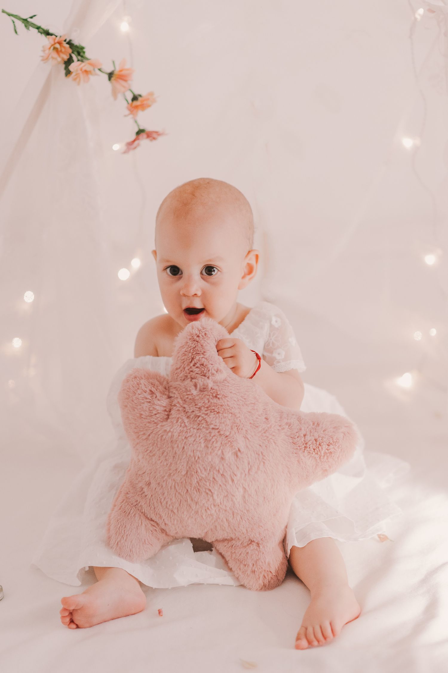 Baby in diaper playing with pink plush toy on white backdrop with twinkling lights.