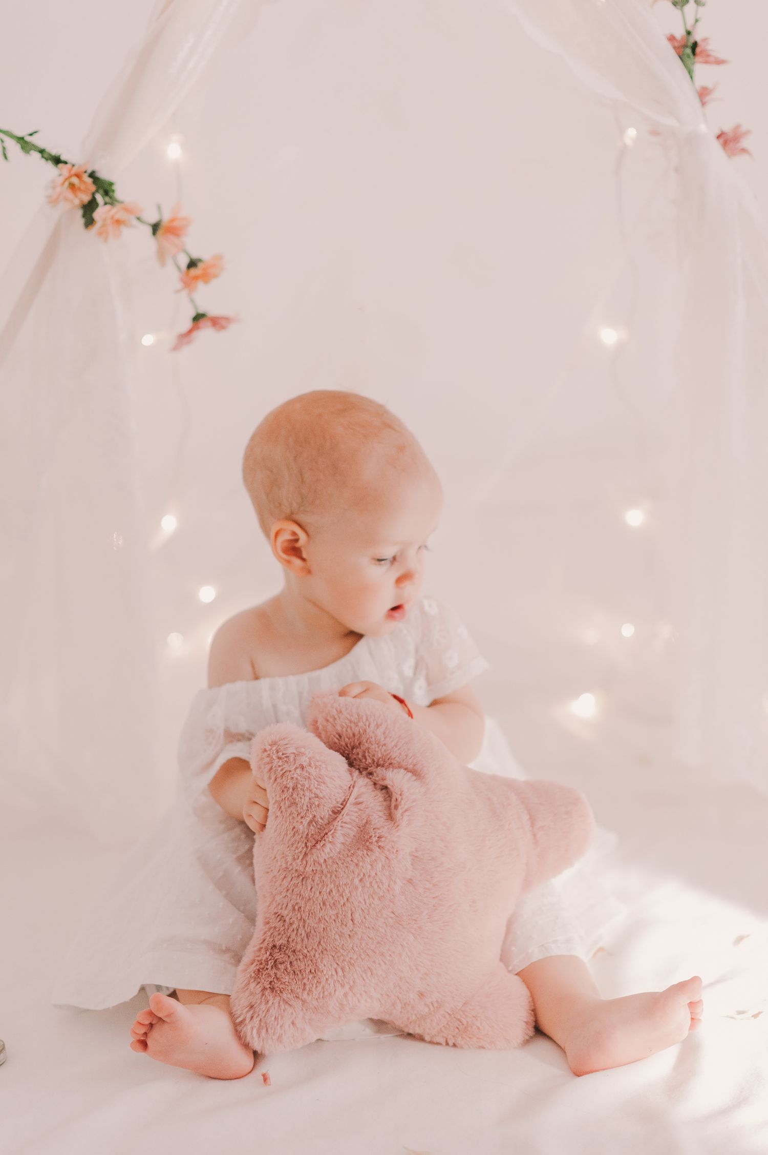Baby sitting on white blanket with pink plush toy and delicate floral decorations.