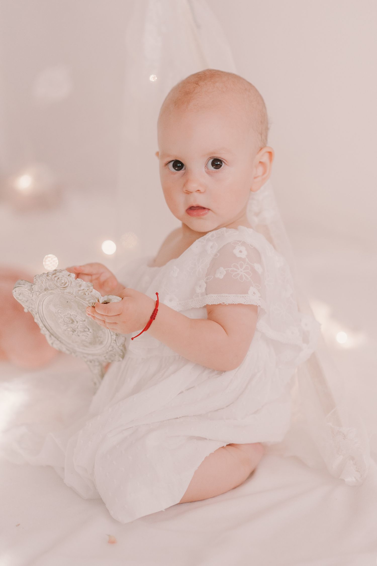 Baby in white dress playing on soft white backdrop with sparkly lighting effects.