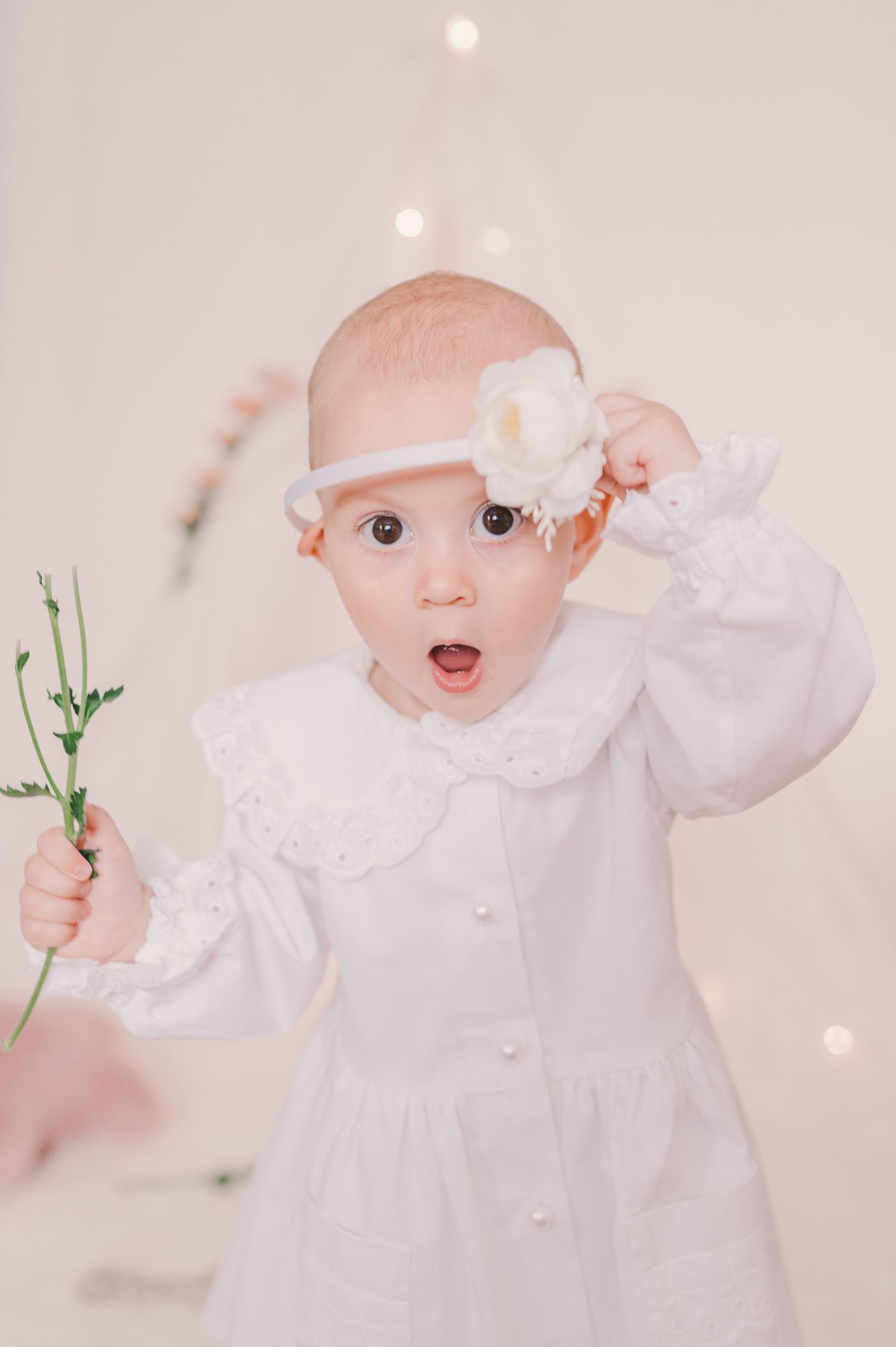 Baby wearing white dress reaching for delicate floral stems against light background.
