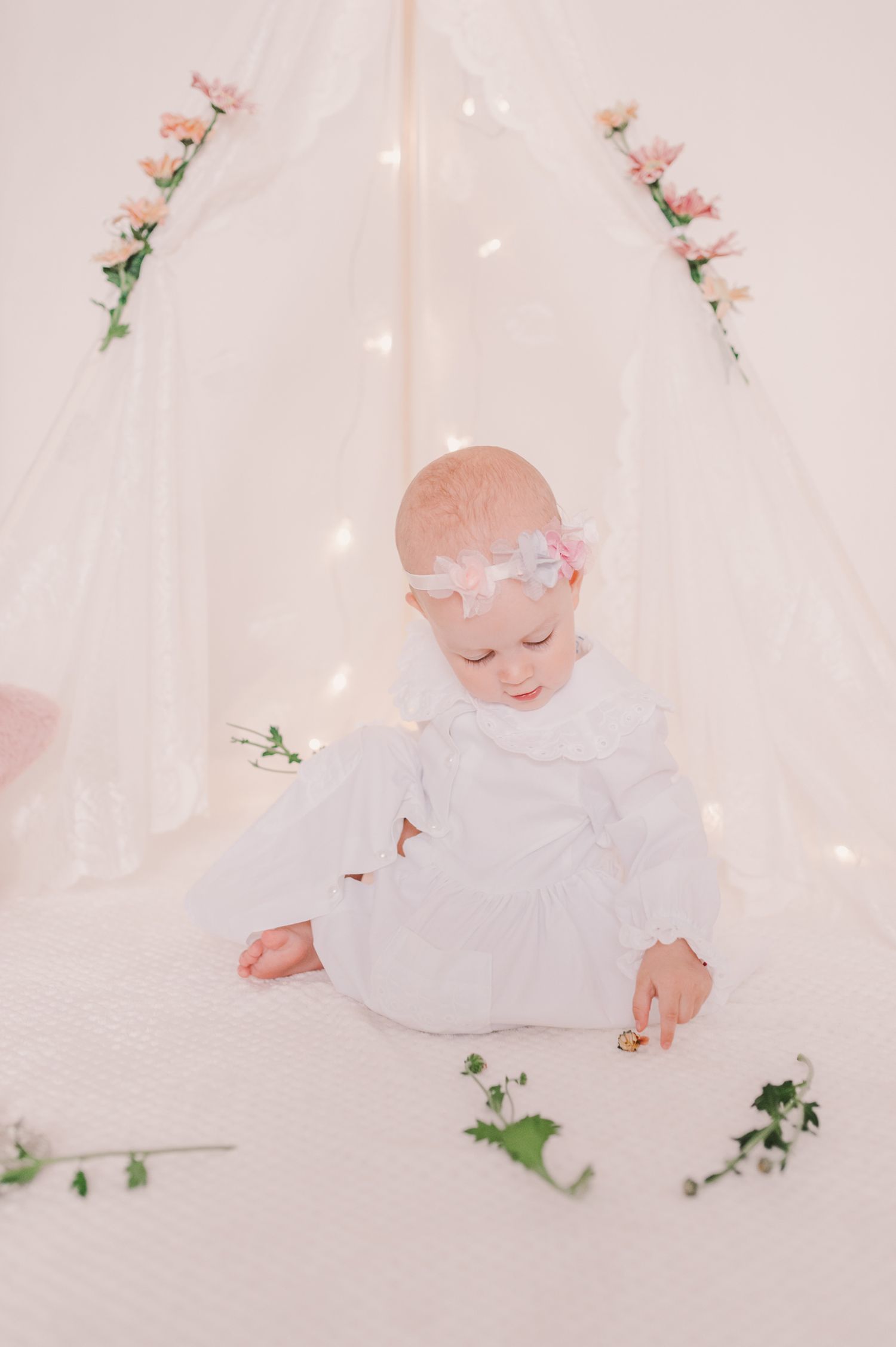 Baby in white dress lying on white backdrop surrounded by scattered flower petals.