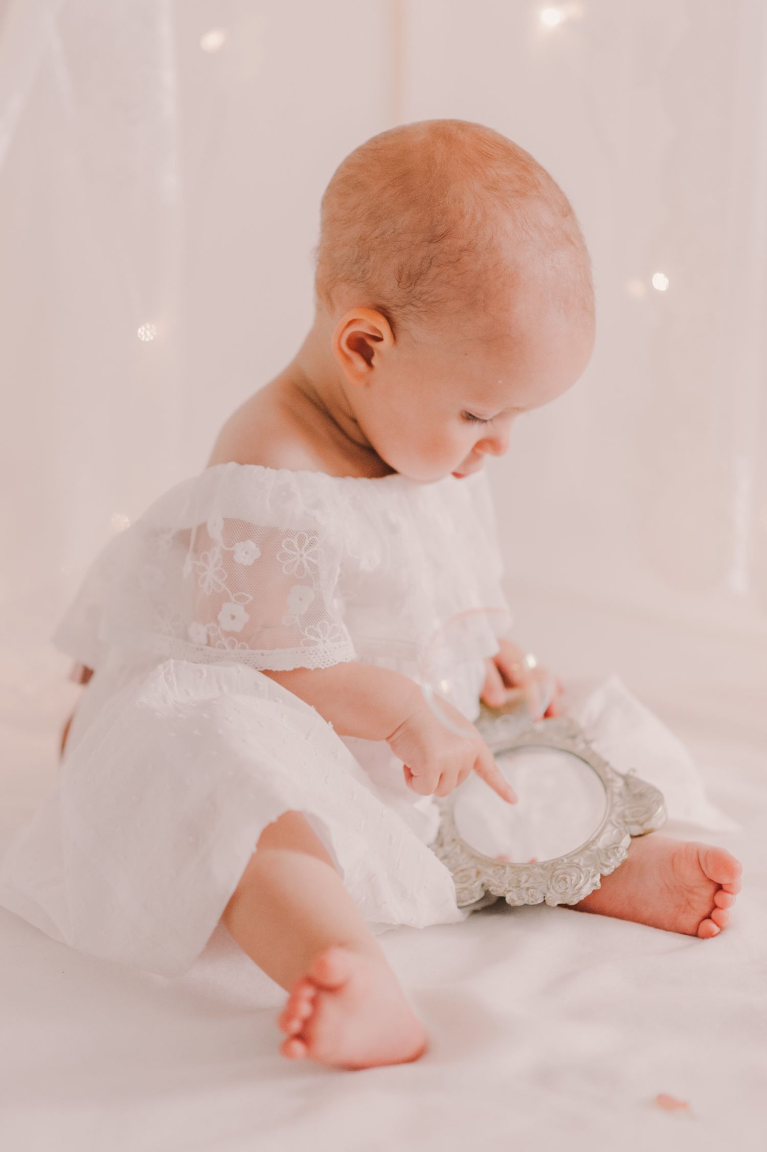 Baby in white lace dress sitting on light backdrop examining decorative mirror.
