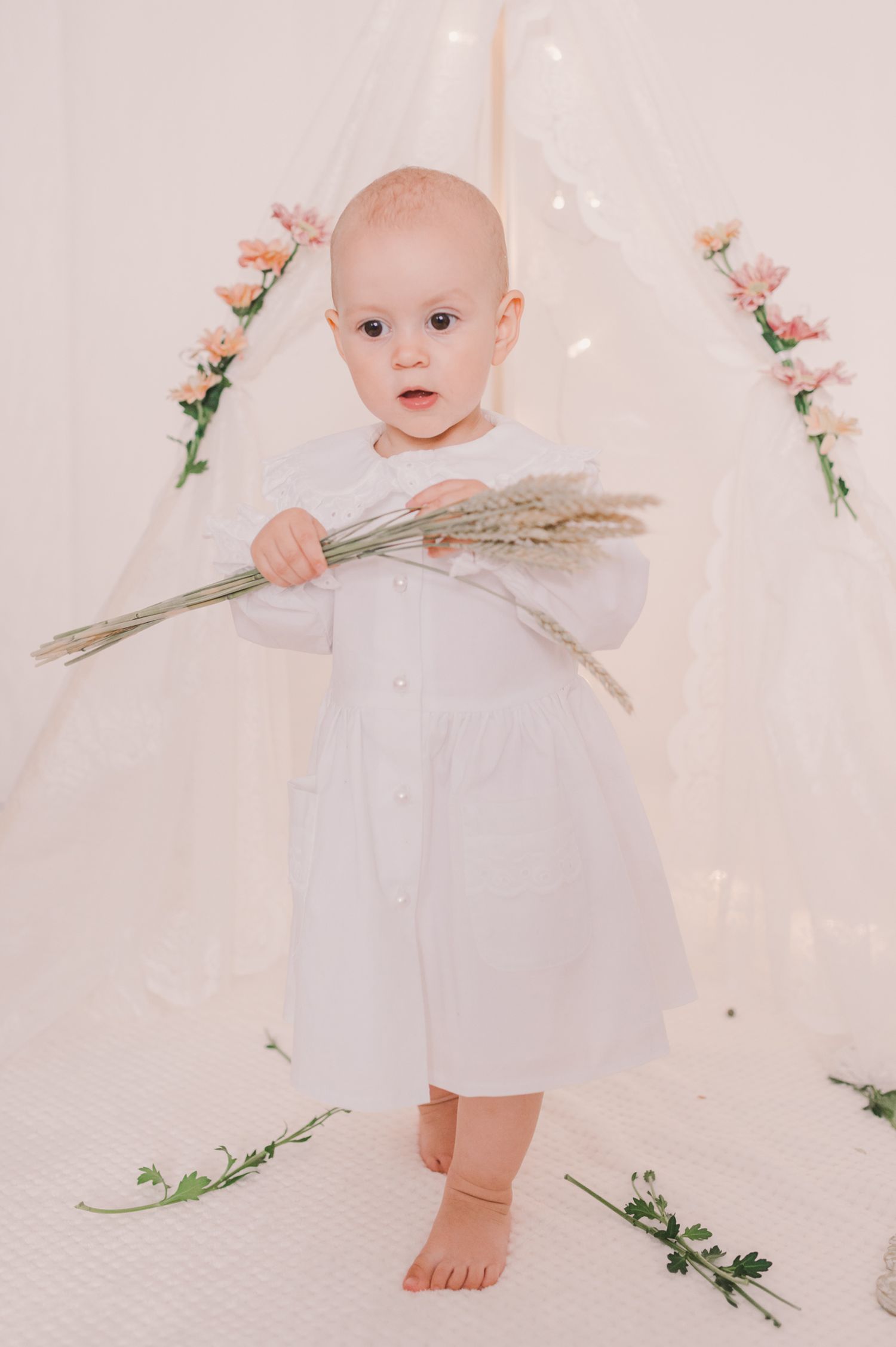Baby in white dress holding dried flowers against ethereal white background.