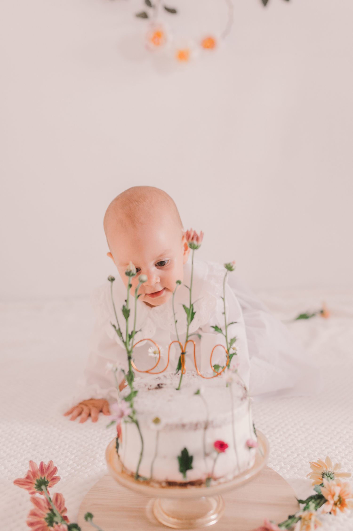 Baby in white dress sits behind delicate floral decorations and glass dome in dreamy pastel setting.